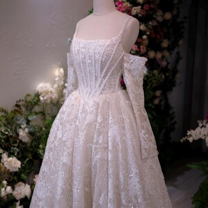 K&ouml;nnte beinhalten: Wei&szlig;es Hochzeitskleid mit quadratischem Ausschnitt und schulterfreien &Auml;rmeln. Das Kleid ist mit aufwendiger Spitze und funkelnden Verzierungen verziert. Eine goldene Krone sitzt auf dem Kopf der Schaufensterpuppe. Das Kleid wird vor einem floralen Hintergrund pr&auml;sentiert.