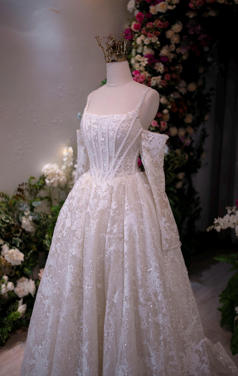 K&ouml;nnte beinhalten: Wei&szlig;es Hochzeitskleid mit quadratischem Ausschnitt und schulterfreien &Auml;rmeln. Das Kleid ist mit aufwendiger Spitze und funkelnden Verzierungen verziert. Eine goldene Krone sitzt auf dem Kopf der Schaufensterpuppe. Das Kleid wird vor einem floralen Hintergrund pr&auml;sentiert.