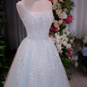 May include: A light blue formal gown on a mannequin, adorned with intricate beadwork and floral embellishments. The dress features a square neckline, spaghetti straps, and a full skirt. A gold crown sits atop the mannequin's head. The background includes a floral arrangement.