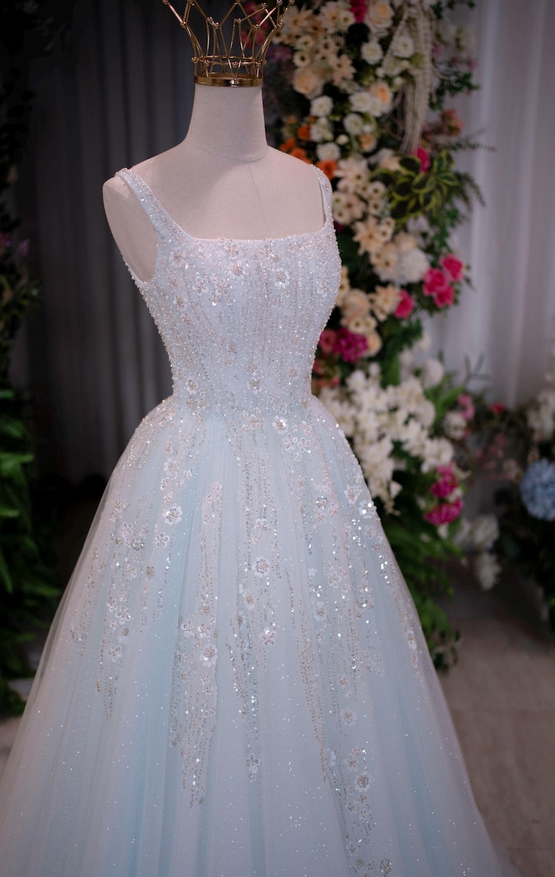 May include: A light blue formal gown on a mannequin, adorned with intricate beadwork and floral embellishments. The dress features a square neckline, spaghetti straps, and a full skirt. A gold crown sits atop the mannequin's head. The background includes a floral arrangement.