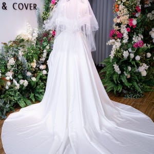 May include: A white bridal gown with a long train and a veil. The dress is displayed on a mannequin in front of a floral arch. The words "MY & COVER" are visible on the wall.