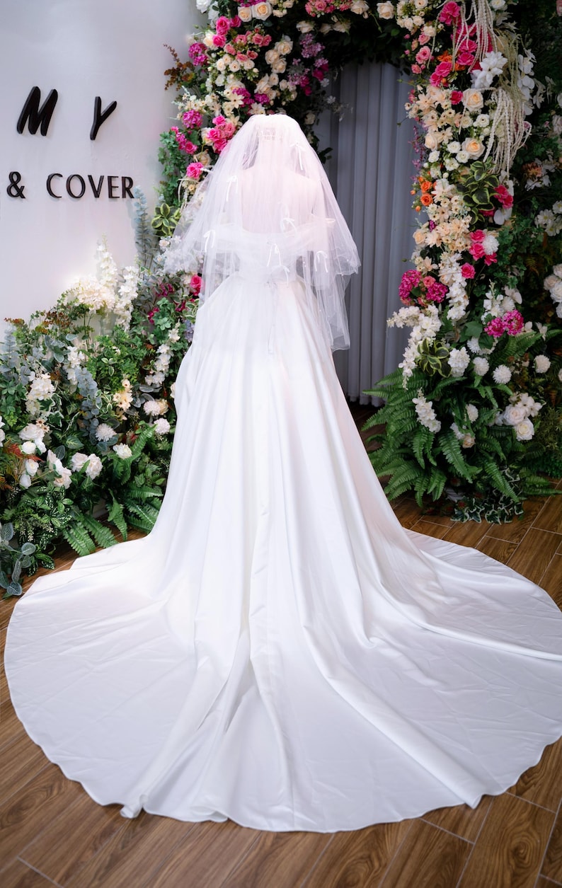 May include: A white bridal gown with a long train and a veil. The dress is displayed on a mannequin in front of a floral arch. The words "MY & COVER" are visible on the wall.