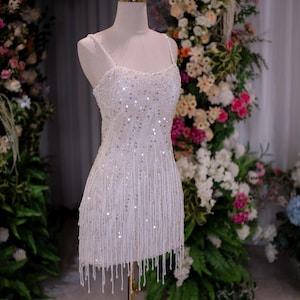 May include: A white, beaded mini dress with spaghetti straps and a fringe hemline. The dress is displayed on a mannequin with a gold crown, set against a backdrop of flowers. The dress is embellished with sequins and hanging beads.