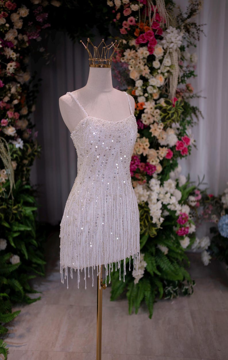 May include: A white, beaded mini dress with spaghetti straps and a fringe hemline. The dress is displayed on a mannequin with a gold crown, set against a backdrop of flowers. The dress is embellished with sequins and hanging beads.