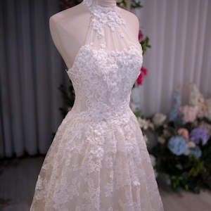 May include: A white sleeveless dress featuring a high halter neckline with intricate floral lace appliqu&eacute;s over a sheer mesh bodice. The skirt is layered with matching white lace embroidery that extends from the waist down to the hemline.
