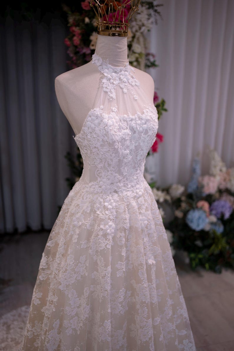 May include: A white sleeveless dress featuring a high halter neckline with intricate floral lace appliqu&eacute;s over a sheer mesh bodice. The skirt is layered with matching white lace embroidery that extends from the waist down to the hemline.