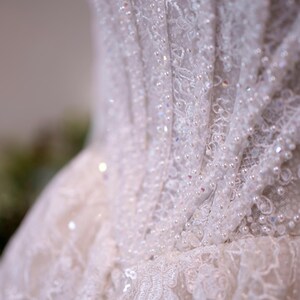 K&ouml;nnte beinhalten: Nahaufnahme eines wei&szlig;en Brautkleid-Oberteils. Das Kleid weist aufwendige Spitzendetails auf, die mit Pailletten und kleinen Perlen verziert sind. Der Stoff ist geschichtet und strukturiert, wodurch ein zartes und elegantes Aussehen entsteht.