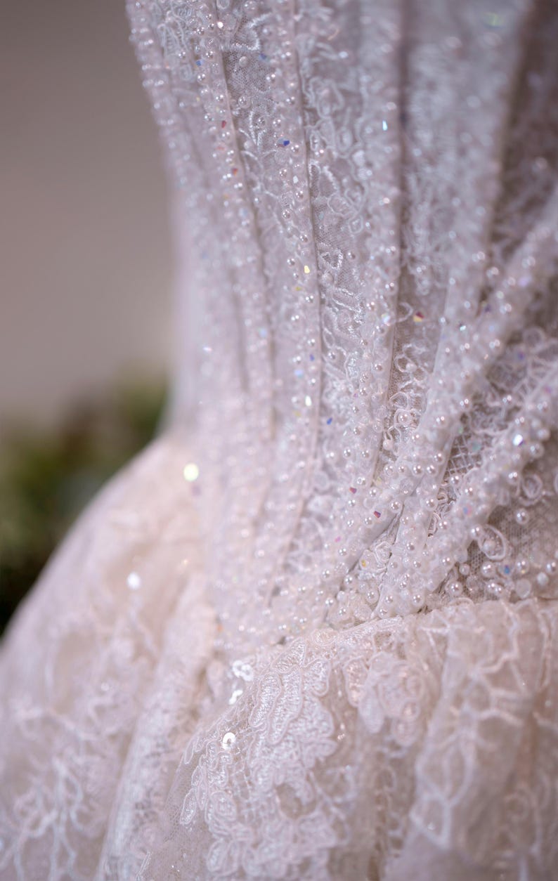 K&ouml;nnte beinhalten: Nahaufnahme eines wei&szlig;en Brautkleid-Oberteils. Das Kleid weist aufwendige Spitzendetails auf, die mit Pailletten und kleinen Perlen verziert sind. Der Stoff ist geschichtet und strukturiert, wodurch ein zartes und elegantes Aussehen entsteht.