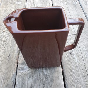 May include: A brown ceramic pitcher with a square body and a handle. The pitcher is sitting on a wooden surface.