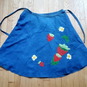 May include: Blue denim apron with a strawberry and flower applique design. The apron features a wrap-around style with long ties. The strawberries are red with white polka dots and green leaves, and the flowers are white with yellow centers.