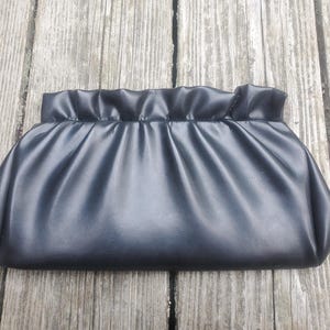 May include: A black clutch purse with a gathered top. The rectangular bag is made of a smooth, dark material and has a simple, elegant design. The purse is approximately 12 inches wide.