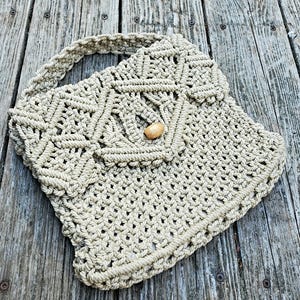 May include: A beige macrame handbag with a woven handle and flap closure. The bag features a geometric pattern and a wooden button clasp. The bag is made of a thick, woven material and is suitable for carrying everyday items.