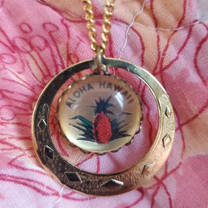 以下が含まれることがあります： パイナップルと「アロハハワイ」の文字が描かれたガラスの円形の中に、丸いペンダントが付いたゴールドネックレスです。