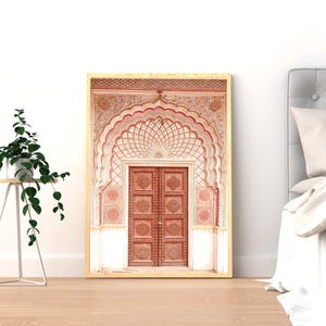 May include: Framed print of an ornate, arched doorway with intricate carvings and a warm color palette of red, pink, and cream. The wooden door features detailed panels. The artwork is displayed in a room with a plant and a bed.
