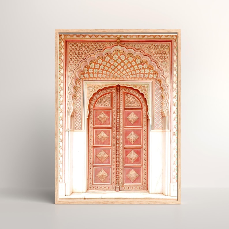 May include: A framed print of an ornate, coral-colored door with intricate carvings. The door is framed by a detailed archway and white pillars, all within a light wood frame. The design evokes a sense of traditional architecture and cultural heritage.