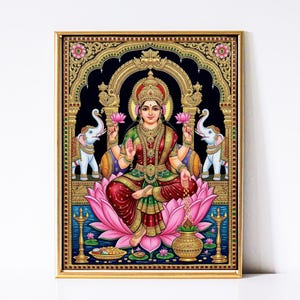Puede incluir: Una pintura enmarcada y colorida de una deidad sentada, adornada con joyas de oro y un sari rojo y verde. La deidad está sentada sobre una flor de loto rosa, sosteniendo flores de loto y un rosario. Dos elefantes blancos flanquean a la deidad, y el fondo es negro.