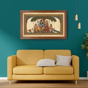 Puede incluir: Una obra de arte enmarcada que representa a una familia hind&uacute; bajo un &aacute;rbol. La obra presenta colores vibrantes y detalles intrincados, con figuras sentadas en una roca. Tambi&eacute;n hay un pavo real y una vaca. El marco es de color marr&oacute;n oscuro.