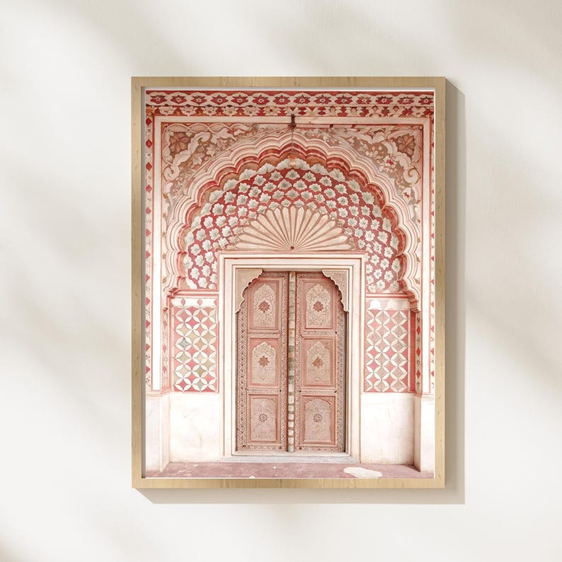 May include: Framed print of an ornate, arched doorway with intricate floral and geometric patterns in shades of red, pink, and white. The wooden door is detailed with carvings. The artwork is set against a white wall.
