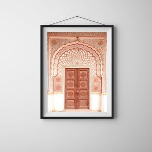 May include: Framed print of an ornate, arched doorway with a carved wooden door. The doorway features intricate patterns in shades of red, cream, and brown. The artwork is displayed in a black frame, hanging against a white wall.