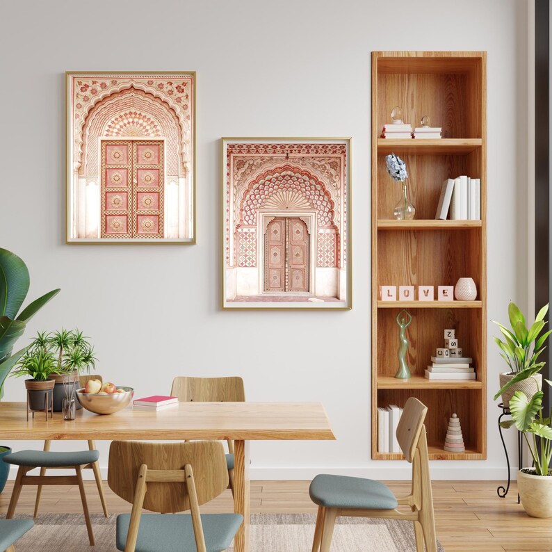 May include: Two framed prints of ornate archways with wooden doors in a room. The prints have gold frames and feature intricate patterns in shades of red and cream. A wooden bookshelf with books and decor is to the right.