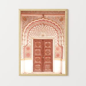 May include: A framed print featuring a detailed architectural facade. The image showcases a carved wooden door within an arched entryway, adorned with intricate patterns in shades of red, pink, and cream. The frame is a light gold color.
