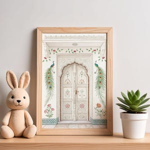May include: A framed print of a white ornate door with floral and peacock accents. The artwork is in a light wood frame and is displayed on a wooden surface. A plush bunny and a potted succulent are next to the framed print.