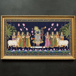 May include: A framed painting depicting a religious scene with figures in traditional attire. The artwork features a central deity surrounded by women, cows, and lotus flowers, all set against a dark blue background. The frame is gold-colored.