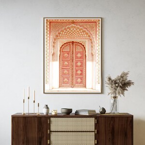 May include: Framed print of a detailed, ornate red door with an arched top. The door is set within a decorative frame with intricate patterns in shades of red, gold, and white. The print is displayed above a dark wood cabinet.