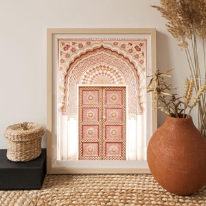 May include: A framed print featuring a detailed illustration of a pink and cream-colored arched doorway. The artwork showcases intricate floral patterns and geometric designs, with a light wooden frame. A woven basket and a terracotta vase are in the foreground.