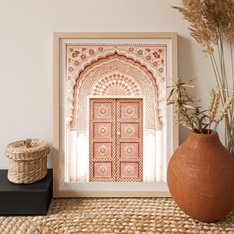 May include: A framed print featuring a detailed illustration of a pink and cream-colored arched doorway. The artwork showcases intricate floral patterns and geometric designs, with a light wooden frame. A woven basket and a terracotta vase are in the foreground.