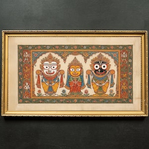 Può includere: Dipinto incorniciato raffigurante tre divinità con grandi occhi e copricapi elaborati. Le figure sono vestite con abiti dai colori vivaci, tra cui giallo, blu e rosso. L'opera d'arte è ambientata su uno sfondo color crema e incorniciata in oro.