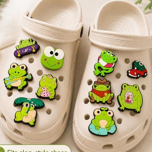 May include: A pair of off-white clog-style shoes decorated with various colourful frog-themed rubber charms. The charms feature frogs in different poses, including riding a skateboard, playing a guitar, wearing a cowboy hat, and holding a milk carton.