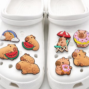 May include: A pair of white clogs decorated with eight colorful rubber charms featuring capybaras in various scenes, including eating watermelon, sitting on a beach chair under an umbrella, wearing a donut, and resting under a rainbow.