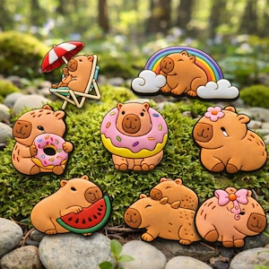 May include: A collection of eight tan capybara-shaped charms featuring various designs: one in a beach chair with a red and white umbrella, one with a rainbow, one holding a pink donut, one wearing a donut, one with a flower, one with a watermelon slice, one with a smaller capybara, and one with a pink bow.