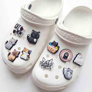 May include: White Crocs sandals adorned with various cat-themed charms. Charms include a black cat, a gray cat, a calico cat, and charms with the words "Cat Mom" and "Pawsitive".