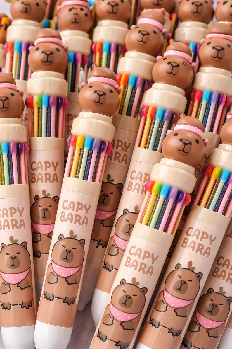 May include: A collection of multi-colored pens with a capybara design. Each pen has a brown capybara head on top and a barrel with the text "CAPY BARA" and a capybara illustration. The pens are arranged in a close-up shot.