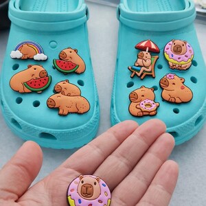 May include: A pair of bright turquoise foam clogs decorated with various capybara-themed shoe charms. The charms feature capybaras with watermelons, rainbows, donuts, and beach chairs. An additional capybara donut charm is held in the foreground.