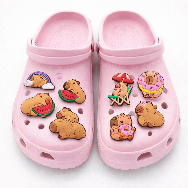May include: A pair of light pink slip-on clogs decorated with various rubber charms featuring capybaras. The charms depict capybaras eating watermelon, sitting on a beach chair under an umbrella, wearing a flower, and holding a donut.