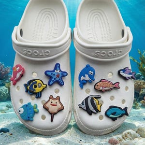 Könnte beinhalten: Ein Paar weiße Clogs, verziert mit bunten Meereskreaturen-Anhängern, darunter Fische, ein Seestern und ein Delfin. Die Clogs befinden sich vor einem Unterwasserhintergrund mit Sand und Wasserpflanzen.