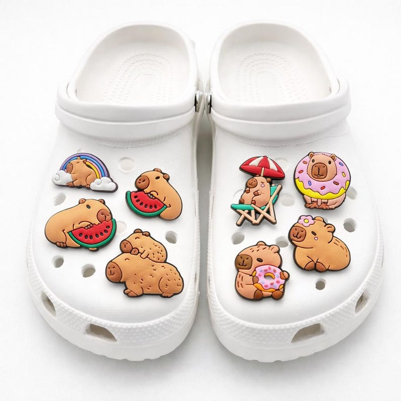 May include: A pair of white clogs decorated with eight colorful rubber charms featuring capybaras in various scenes, including eating watermelon, sitting on a beach chair under an umbrella, wearing a donut, and resting under a rainbow.