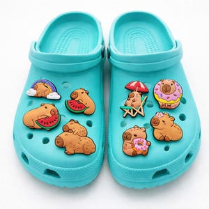May include: A pair of bright turquoise foam clogs decorated with various capybara-themed charms. The charms feature capybaras with rainbows, watermelons, beach chairs, umbrellas, donuts, and flowers, all attached to the holes of the footwear.