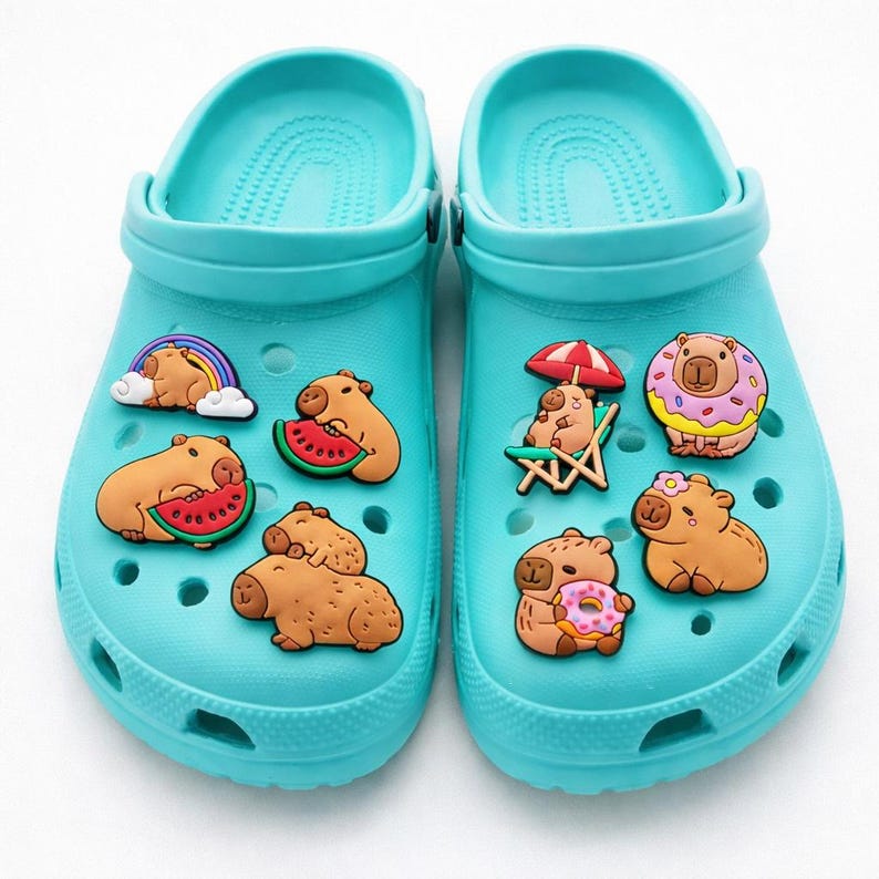 May include: A pair of bright turquoise foam clogs decorated with various capybara-themed charms. The charms feature capybaras with rainbows, watermelons, beach chairs, umbrellas, donuts, and flowers, all attached to the holes of the footwear.