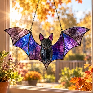May include: A stained glass bat suncatcher featuring purple and blue textured glass panels with black metal framing. The bat is suspended by two thin black chains and displays pointed ears and outstretched wings with a reflective finish.
