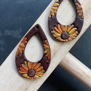 May include: A pair of brown leather teardrop-shaped earrings with a sunflower design. The sunflowers are painted in shades of orange, yellow, and white.