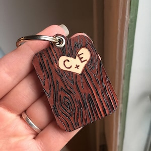 May include: A wooden keychain with a heart-shaped design and the initials "C+E" engraved in the center. The keychain has a wood grain pattern and a brown color.