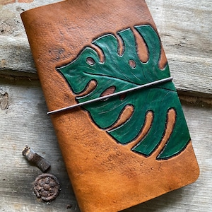 May include: A brown leather journal with a hand-painted green monstera leaf design. The journal is closed with a metal clasp. The leather has a textured appearance, and the leaf is detailed with veins.