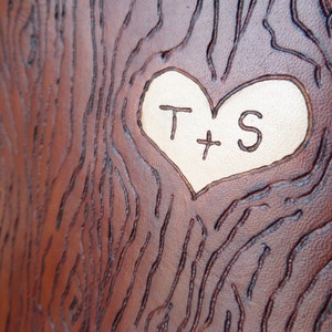 May include: A brown leather surface with a carved heart shape. The heart is filled with a lighter brown leather and has the initials "T + S" carved into it.