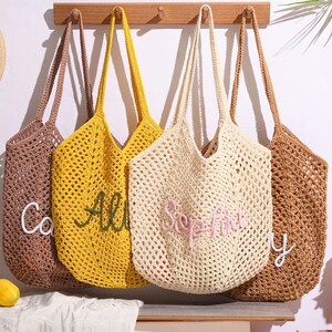 May include: Four open-weave crochet tote bags in brown, yellow, cream, and tan hang from a wooden rack. Each bag has a long strap and a personalized name embroidered on the front in a contrasting color. A straw hat with a black ribbon is visible on the left.