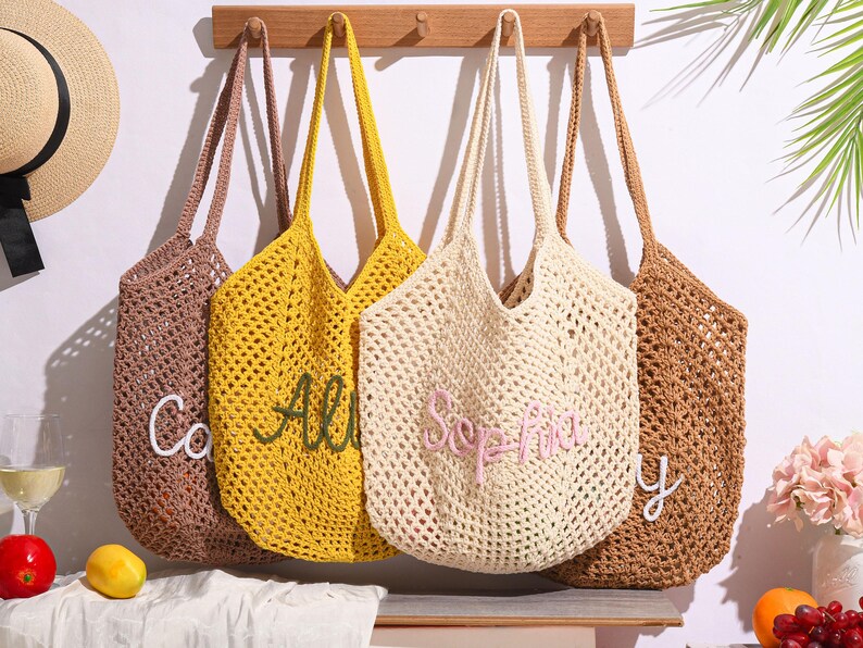 May include: Four open-weave crochet tote bags in brown, yellow, cream, and tan hang from a wooden rack. Each bag has a long strap and a personalized name embroidered on the front in a contrasting color. A straw hat with a black ribbon is visible on the left.