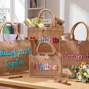 May include: Four rectangular tan jute tote bags with brown handles and button closures. The bags feature colorful, raised, soft-textured lettering spelling out Class of 2026, Sophie, Mrs. White, Whitney, and Mrs. Smith, with a small flower accent.